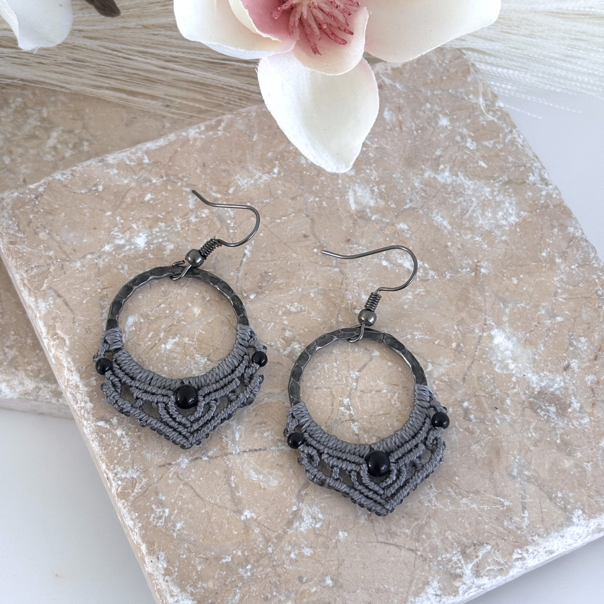 Pair of handmade macrame hoop earrings with 25mm hammered gunmetal rings, gray macrame lattice knotwork, 4mm round black onyx center beads, 2mm round black metal accent beads, and gunmetal fishhook ear wires, displayed on travertine tile with magnolia flower.