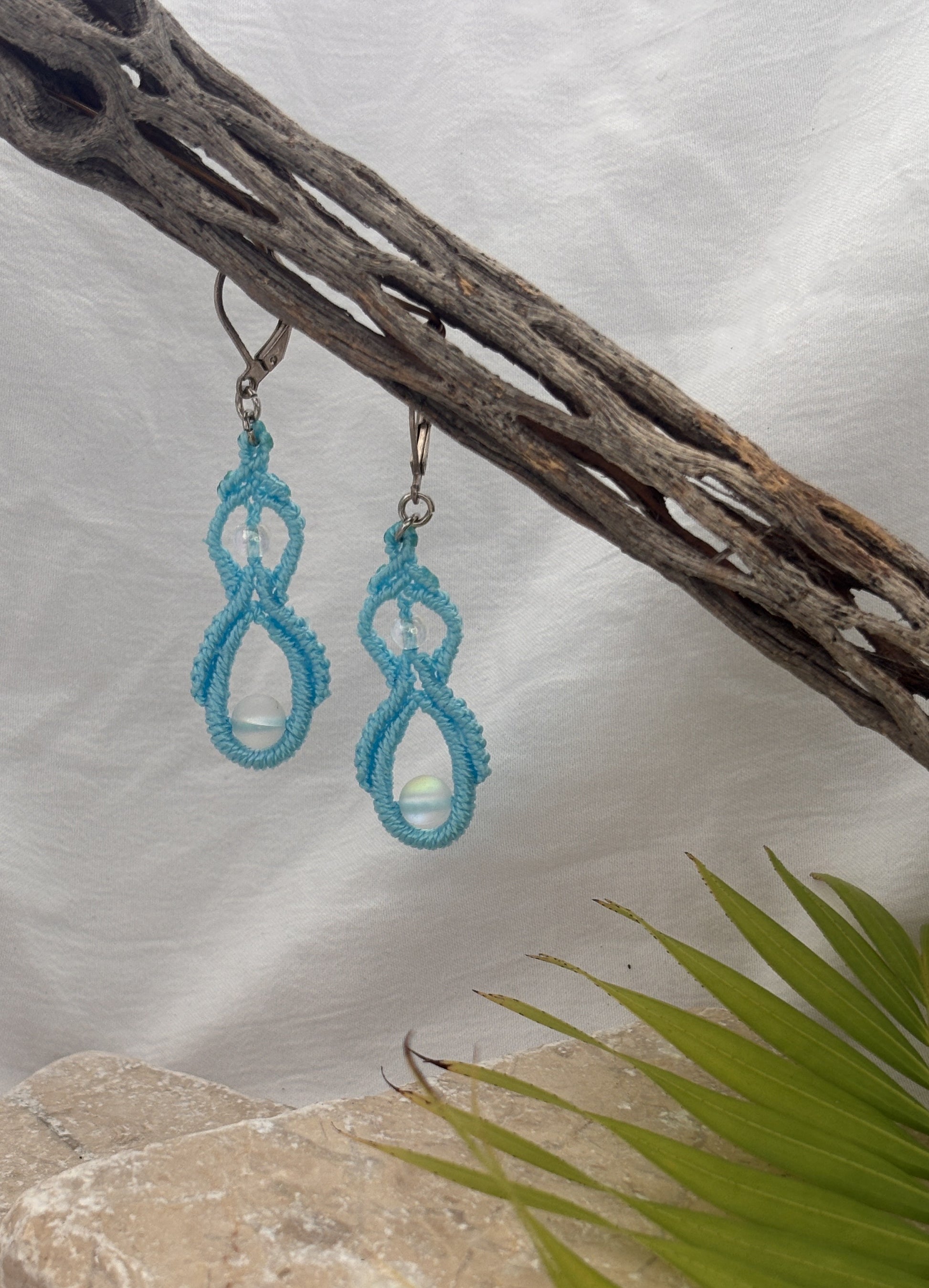 Handmade turquoise macrame double drop earrings with iridescent mermaid glass beads hanging from driftwood with a coastal water and palm backdrop.