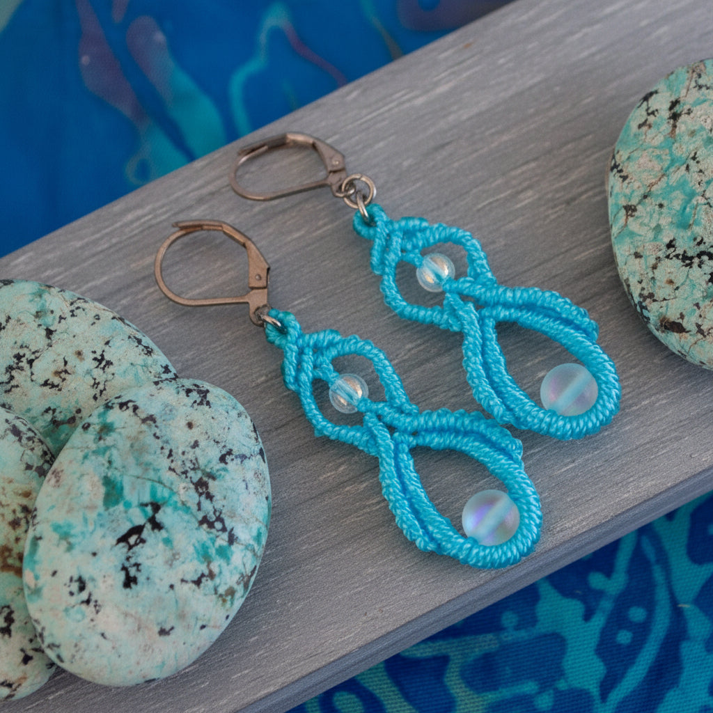 Handmade turquoise macrame double drop earrings with iridescent white mermaid glass beads and silver-tone lever-back closures, displayed on a white wood surface with green speckled stones and a blue background.