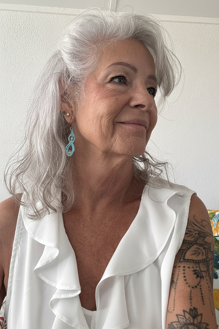 Woman with silver hair wearing handmade turquoise macrame double drop earrings with iridescent mermaid glass beads and silver-tone lever-back closures, styled with a white ruffled top.