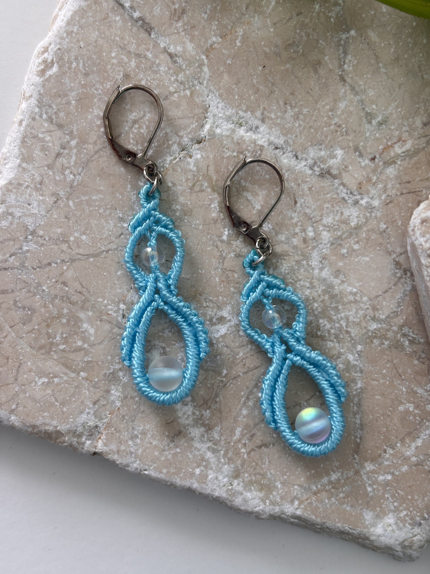 Pair of handmade turquoise macrame double drop earrings with iridescent mermaid glass beads and silver-tone lever-back closures, laid flat on a natural stone surface.