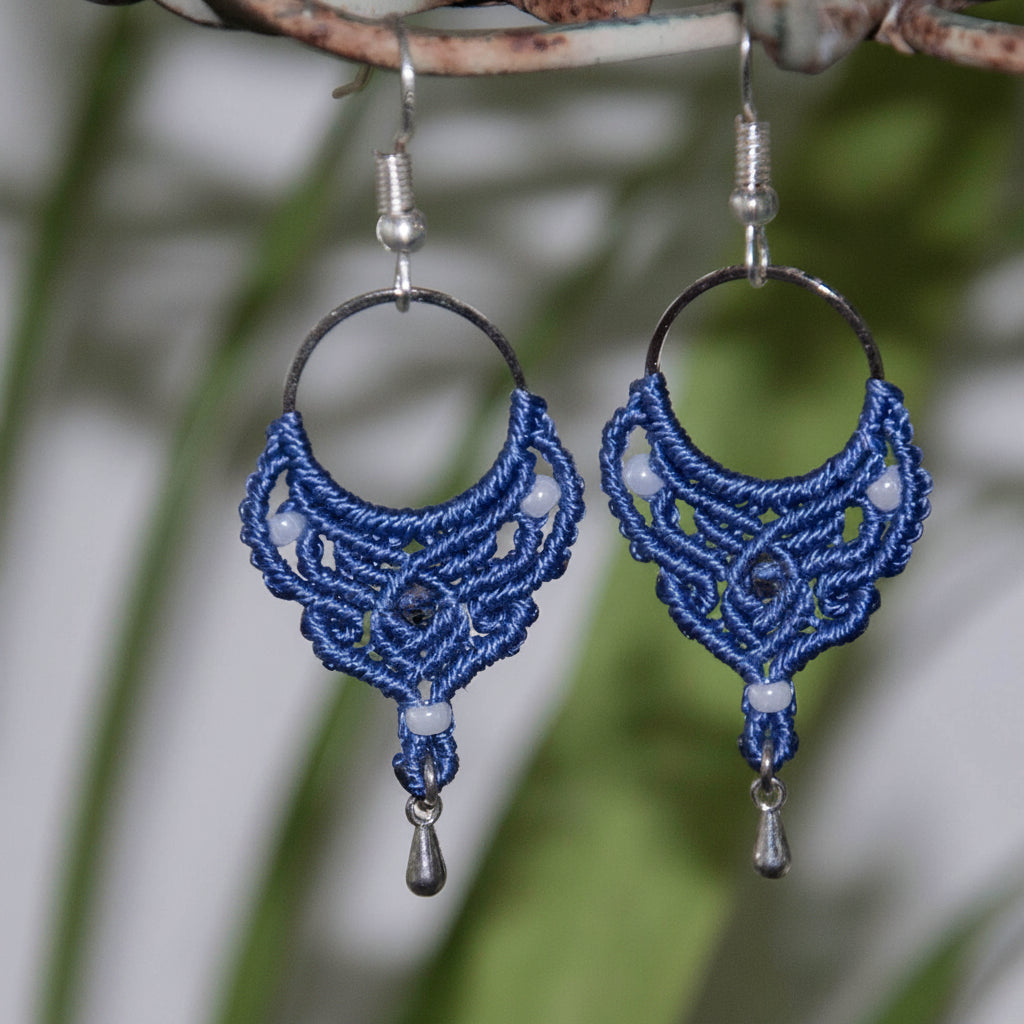 Handmade purple macrame hoop drop earrings with a silver-tone hoop finding, pearlescent white seed beads, faceted purple crystal center bead, and silver-tone waterdrop charms, hanging from a rustic branch with tropical green leaves.