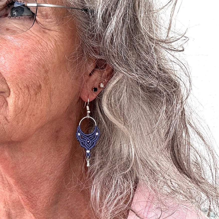 Handmade purple macrame hoop drop earring with silver-tone hoop finding, pearlescent white seed beads, faceted purple crystal center bead, and silver-tone waterdrop charm, shown worn on model with silver curly hair and wire-rimmed glasses.