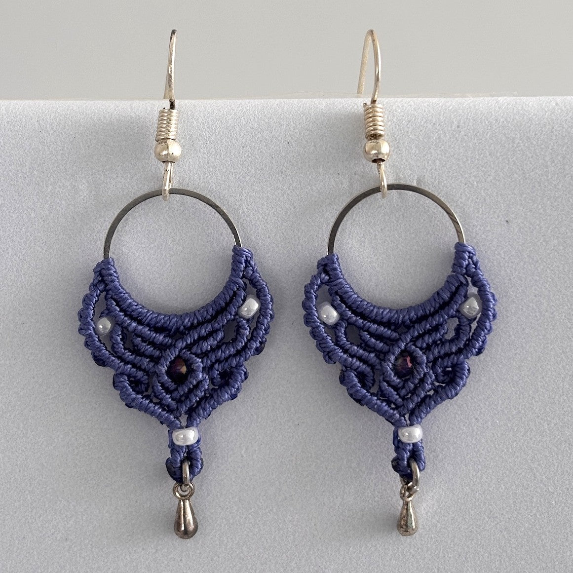 Close-up of handmade purple macrame hoop drop earrings with silver-tone hoop finding, pearlescent white seed beads, faceted purple crystal center bead, and silver-tone waterdrop charms, displayed on a light gray surface.