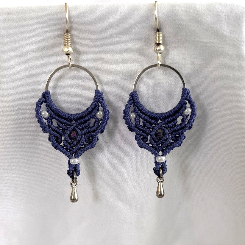Handmade purple macrame hoop drop earrings with silver-tone hoop finding, pearlescent white seed beads, faceted purple crystal center bead, and silver-tone waterdrop charms, laid on draped white fabric.