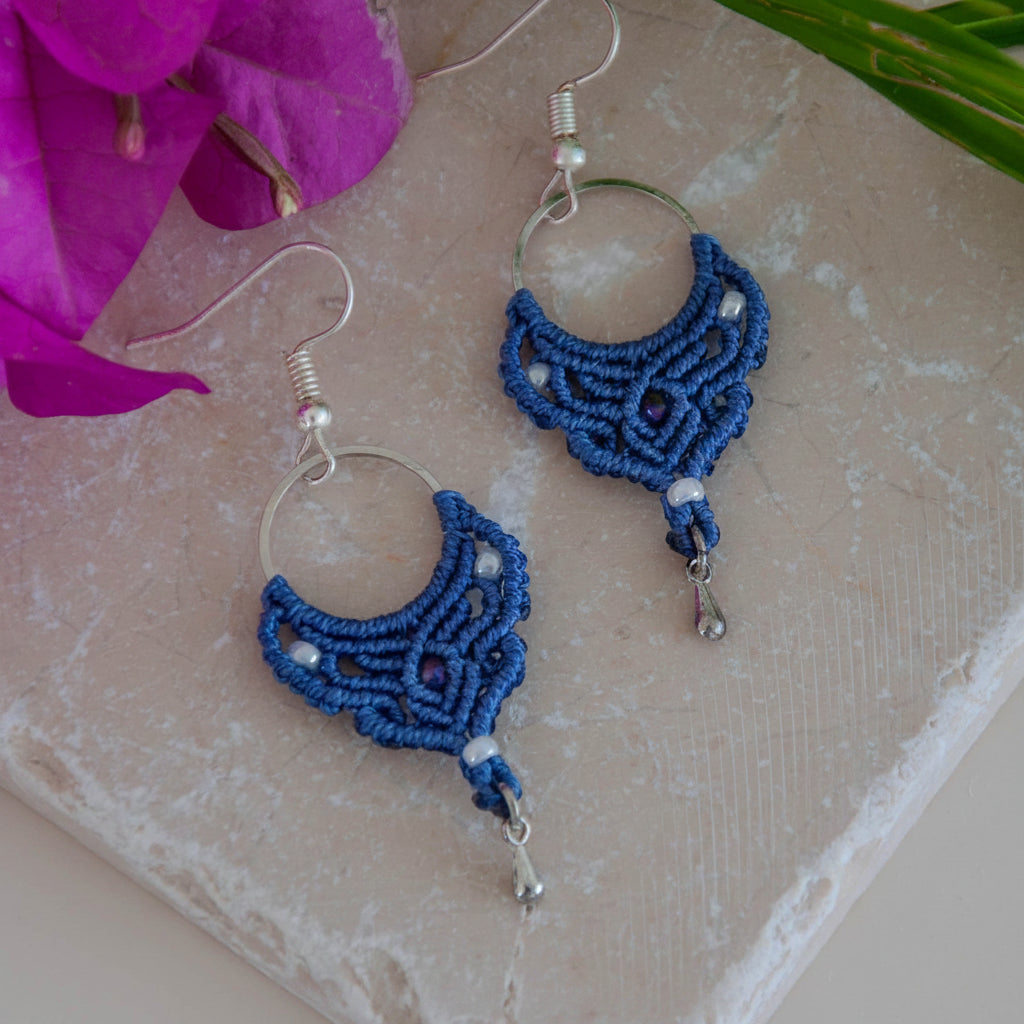 Handmade purple macrame hoop drop earrings with a silver-tone hoop finding, pearlescent white seed beads, faceted purple crystal center bead, and silver-tone waterdrop charms, laid flat on a natural stone surface with magenta bougainvillea flowers.