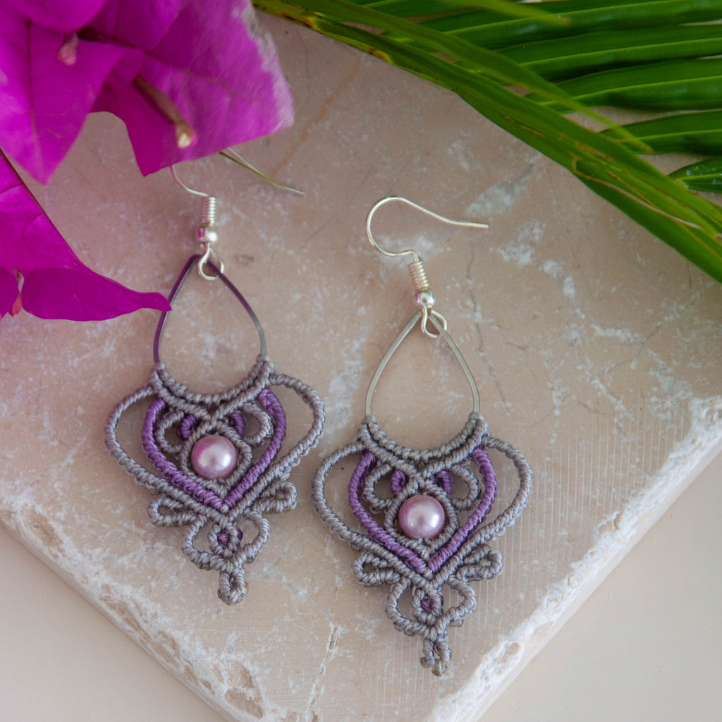 Handmade two-tone silvery gray and purple macrame teardrop drop earrings with an ornate heart-shaped lattice, mauve shell pearl center bead, and silver-tone fish hook wires, laid flat on a natural stone surface with magenta bougainvillea flowers and tropical greenery.