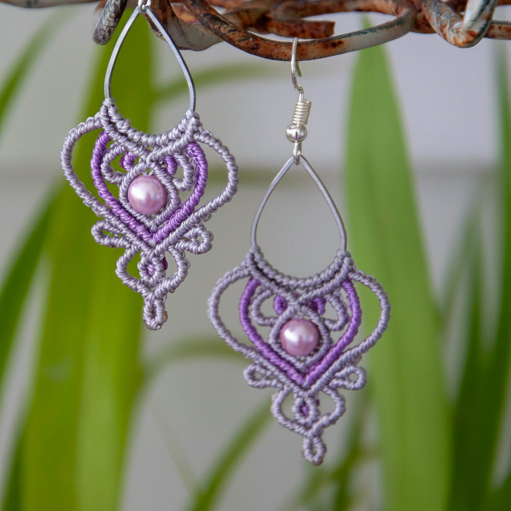Handmade two-tone silvery gray and purple macrame teardrop drop earrings with an ornate heart-shaped lattice, mauve shell pearl center bead, and silver-tone fish hook wires, hanging from a rustic metal stand with tropical green leaves.