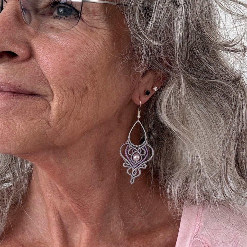Handmade two-tone silvery gray and purple macrame teardrop drop earring with ornate heart-shaped lattice, mauve shell pearl center bead, and silver-tone fish hook wire, shown worn on model with silver curly hair and rimless glasses, pink top.