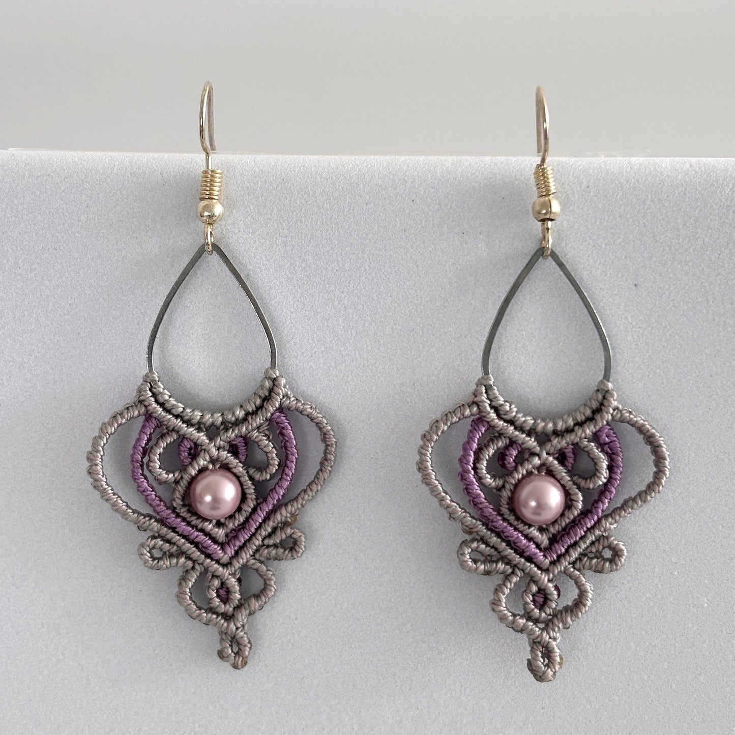 Close-up of handmade two-tone silvery gray and purple macrame teardrop drop earrings with ornate heart-shaped lattice, mauve shell pearl center bead, and silver-tone fish hook wires, displayed on a light gray surface.