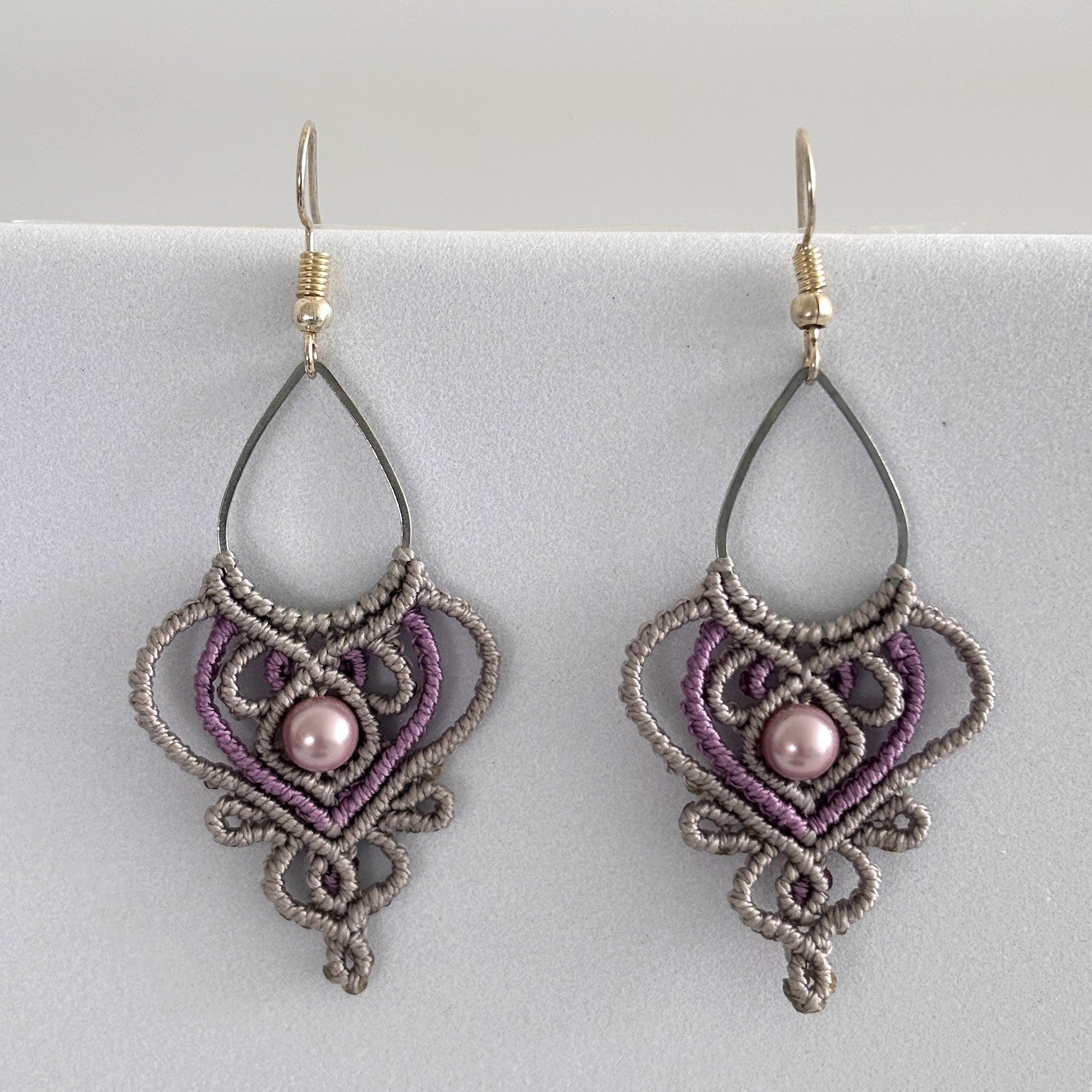 Close-up of handmade two-tone silvery gray and purple macrame teardrop drop earrings with ornate heart-shaped lattice, mauve shell pearl center bead, and silver-tone fish hook wires, displayed on a light gray surface.