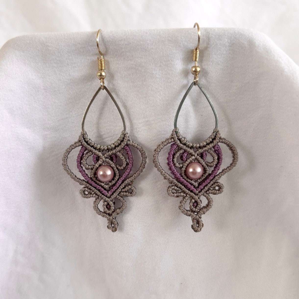 Handmade two-tone silvery gray and purple macrame teardrop drop earrings with ornate heart-shaped lattice, mauve shell pearl center bead, and silver-tone fish hook wires, laid on draped white fabric.
