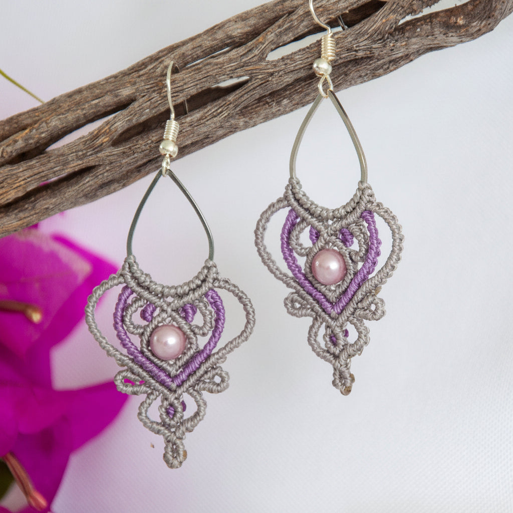 Handmade two-tone silvery gray and purple macrame teardrop drop earrings with an ornate heart-shaped lattice, mauve shell pearl center bead, and silver-tone fish hook wires, hanging from a driftwood branch with magenta bougainvillea flowers.