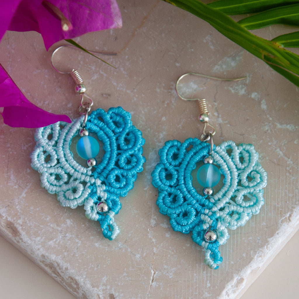 Handmade two-tone turquoise and aqua macrame scroll drop earrings with a turquoise mermaid glass bead center, silver-tone metal bead accents, and silver-tone fish hook wires, laid on a travertine tile with magenta bougainvillea and tropical palm leaves.