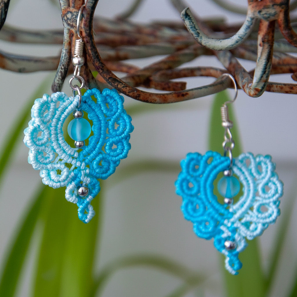 Handmade two-tone turquoise and aqua macrame scroll drop earrings with a turquoise mermaid glass bead center, silver-tone metal bead accents, and silver-tone fish hook wires, hanging from a rustic metal stand with tropical green leaves in the background.