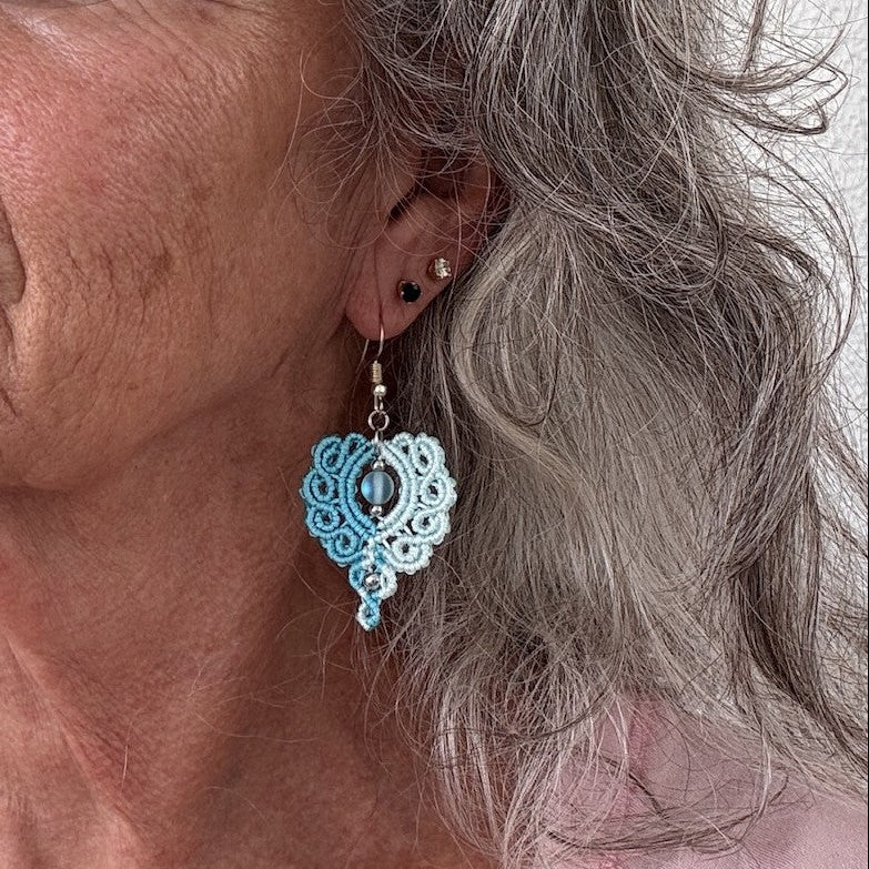 Handmade two-tone turquoise and aqua macrame scroll drop earring with turquoise mermaid glass bead center and silver-tone accents, shown worn on model with silver hair.
