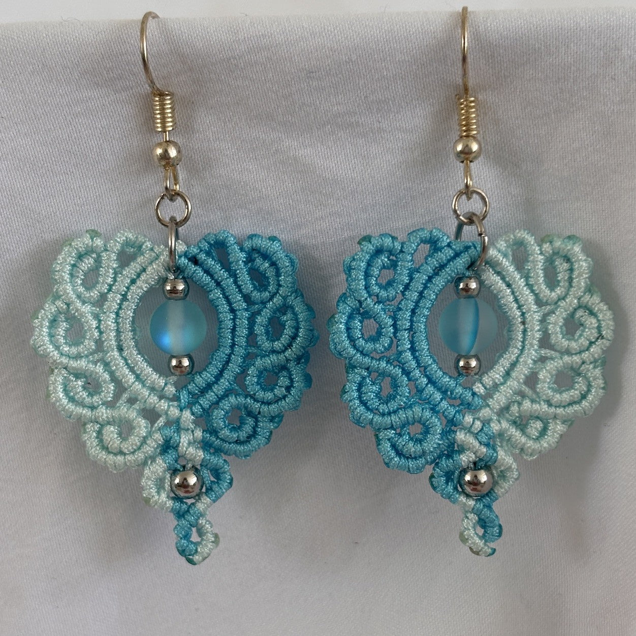 Pair of handmade two-tone turquoise and aqua macrame scroll drop earrings with turquoise mermaid glass bead centers and silver-tone metal bead accents, displayed on white fabric background.
