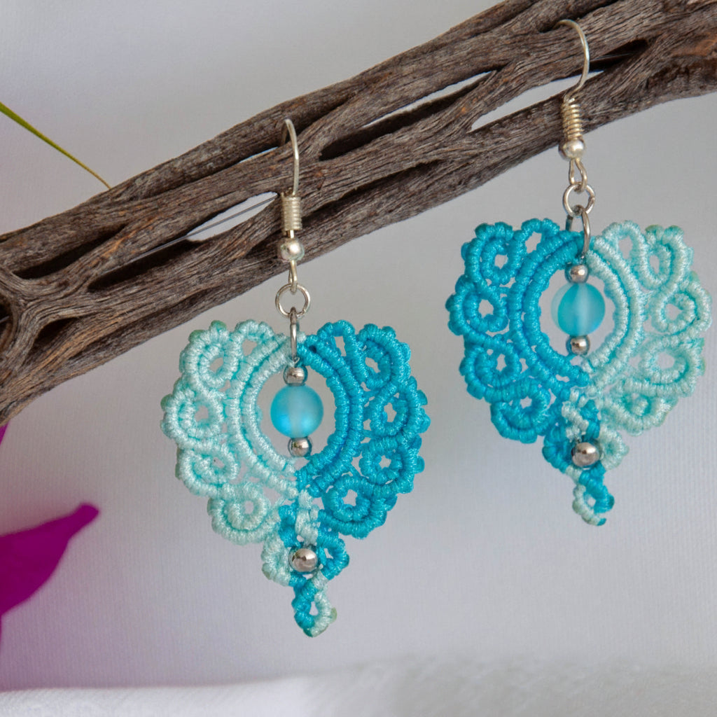 Handmade two-tone turquoise and aqua macrame scroll drop earrings with a turquoise mermaid glass bead center, silver-tone metal bead accents, and silver-tone fish hook wires, hanging from a driftwood branch with magenta bougainvillea against a white background.