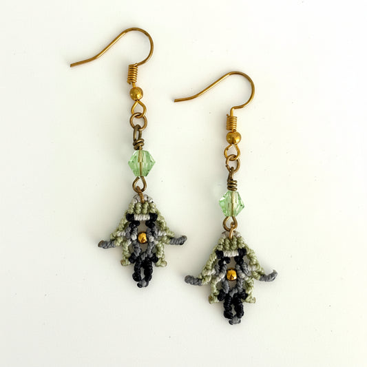 Handmade macrame earrings in light green, black, and silver gray with wire-wrapped light green bicone bead and gold-tone accent, displayed on white background