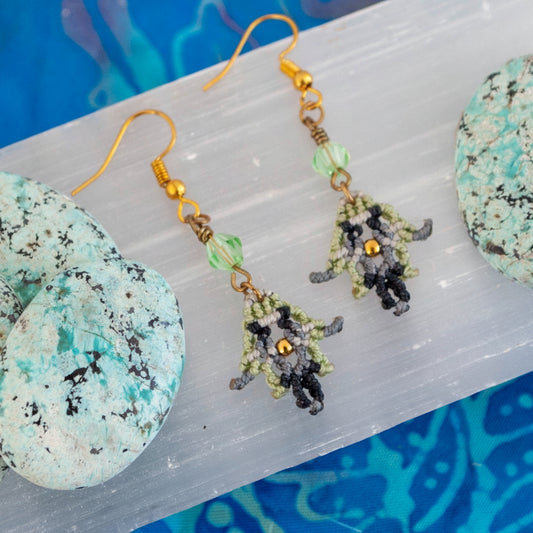 Macrame earrings in light green, black, and silver gray with light green bicone bead, displayed on selenite with teal stones and blue linen background