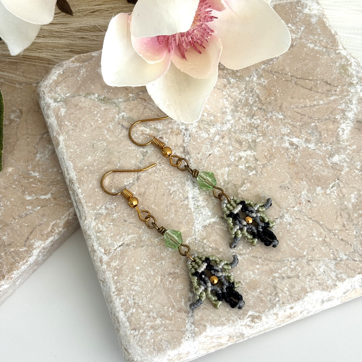 Pair of macrame earrings with light green bicone bead and gold-tone seed bead accent, displayed on travertine tile with magnolia flower