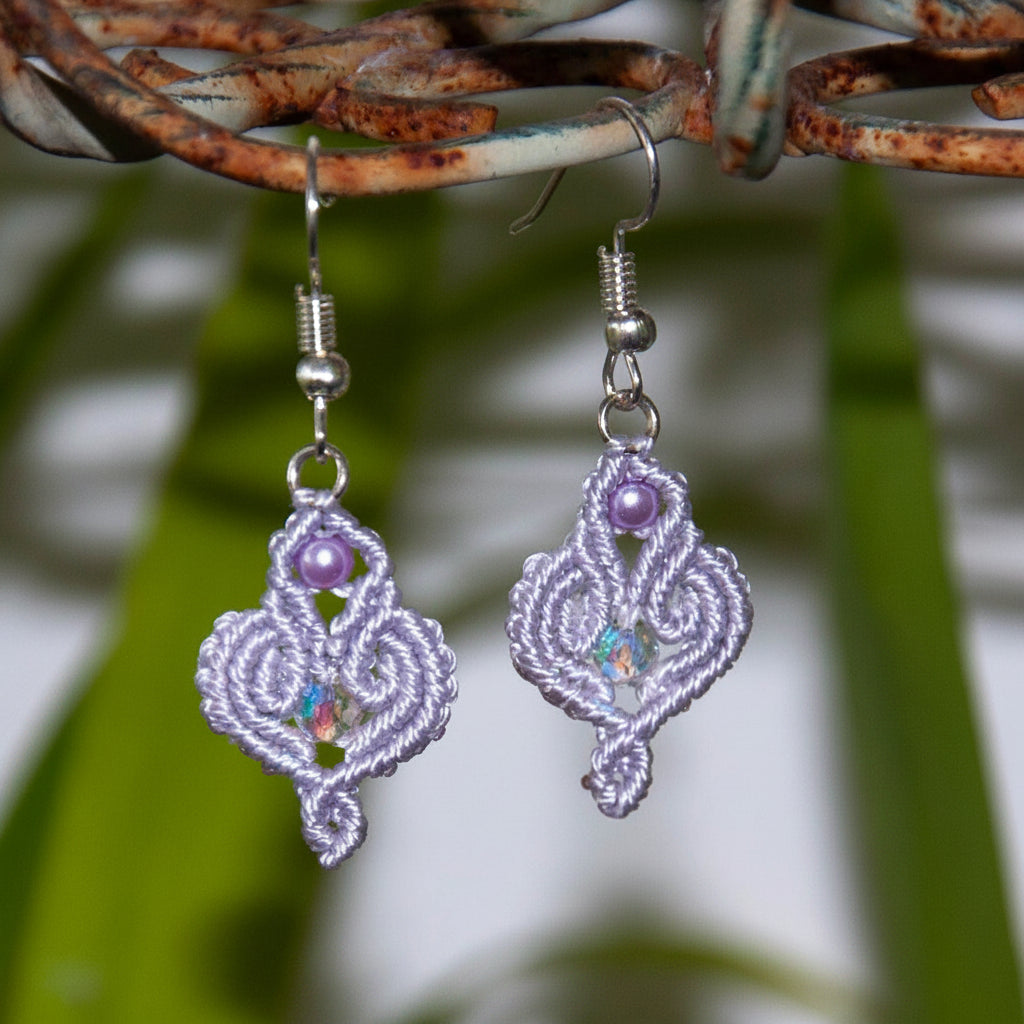 Handmade lavender macrame scroll drop earrings with a lavender glass pearl accent, faceted iridescent crystal center, and silver-tone fish hook wires, hanging from a rustic metal stand with tropical green leaves in the background.