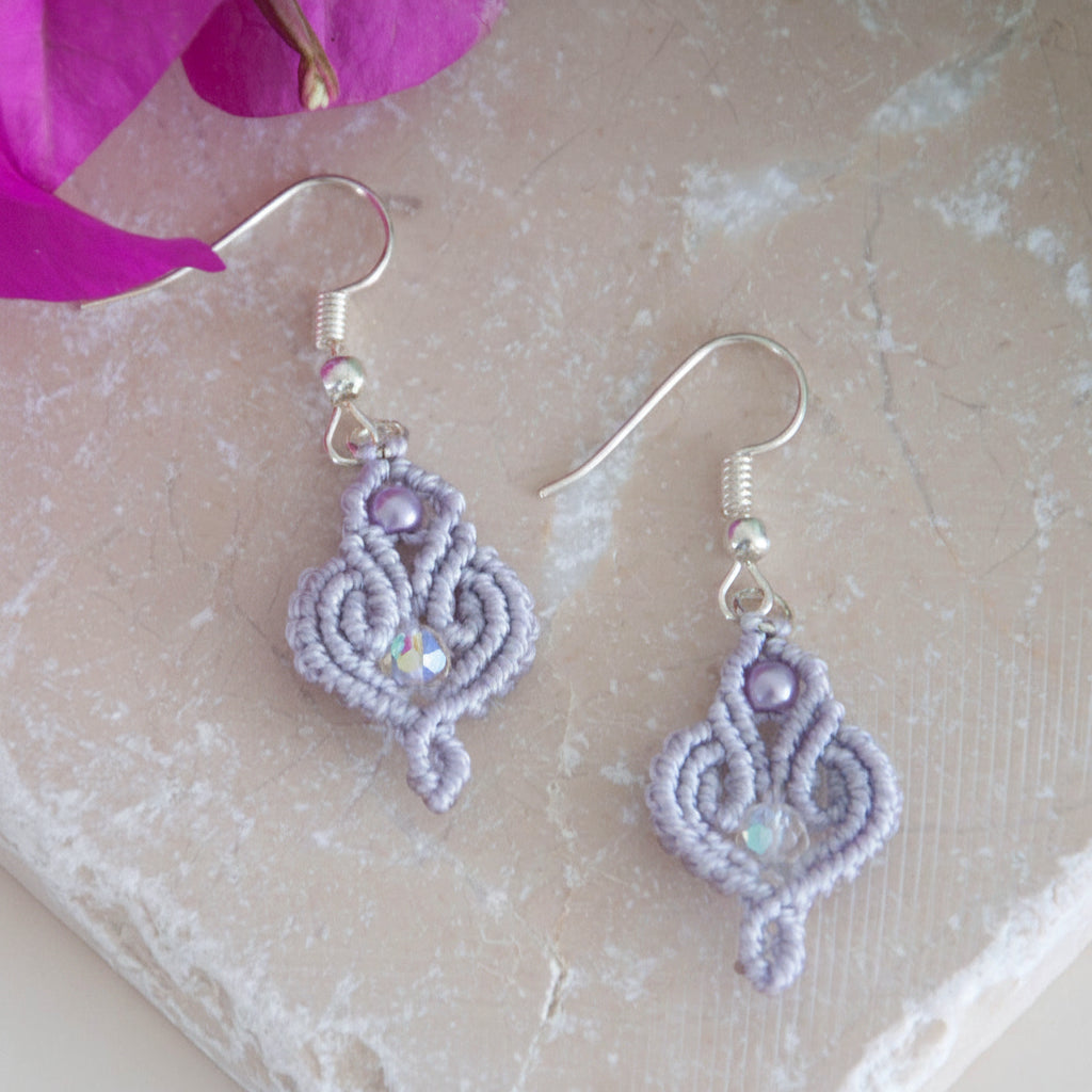 Handmade lavender macrame scroll drop earrings with a lavender glass pearl accent, faceted iridescent crystal center, and silver-tone fish hook wires, laid on a travertine tile with magenta bougainvillea flowers.