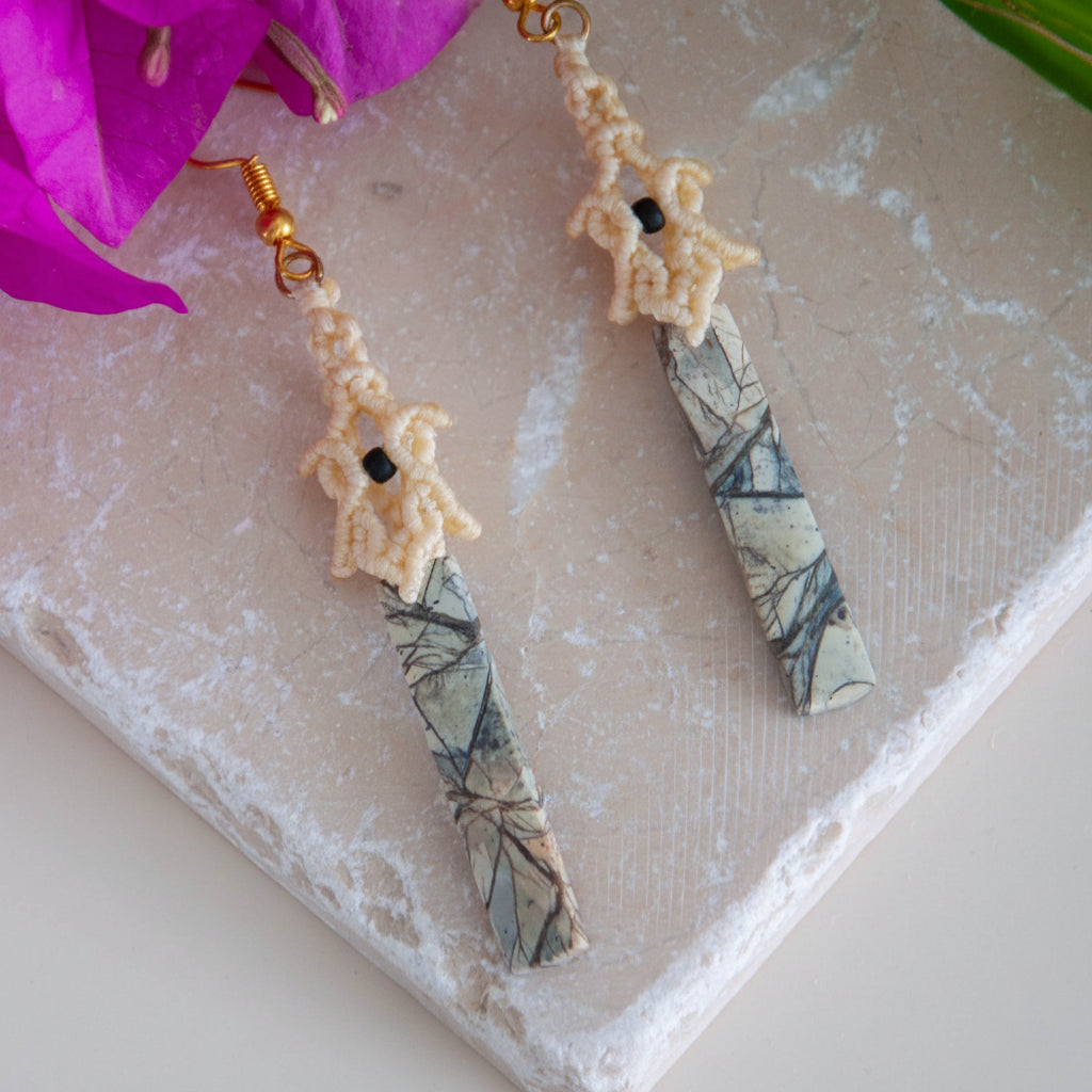 Pair of handmade lace macrame pagoda earrings with maligano jasper stick beads and gold-tone ear wires, displayed on travertine tile with pink bougainvillea