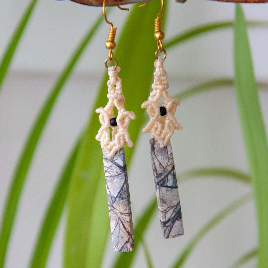 Handmade pale yellow macrame pagoda earrings with maligano jasper stick beads, hanging from driftwood with lush tropical palm leaves in background