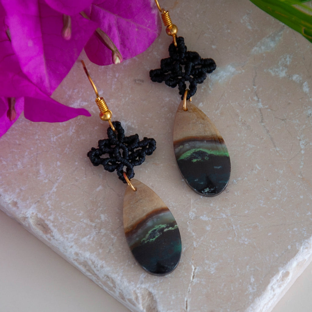 Pair of handmade black macrame drop earrings with diamond-shaped knotwork and green chalcedony teardrop gemstones on gold-tone fish hook wires, laid flat on a natural stone surface with magenta bougainvillea flowers.