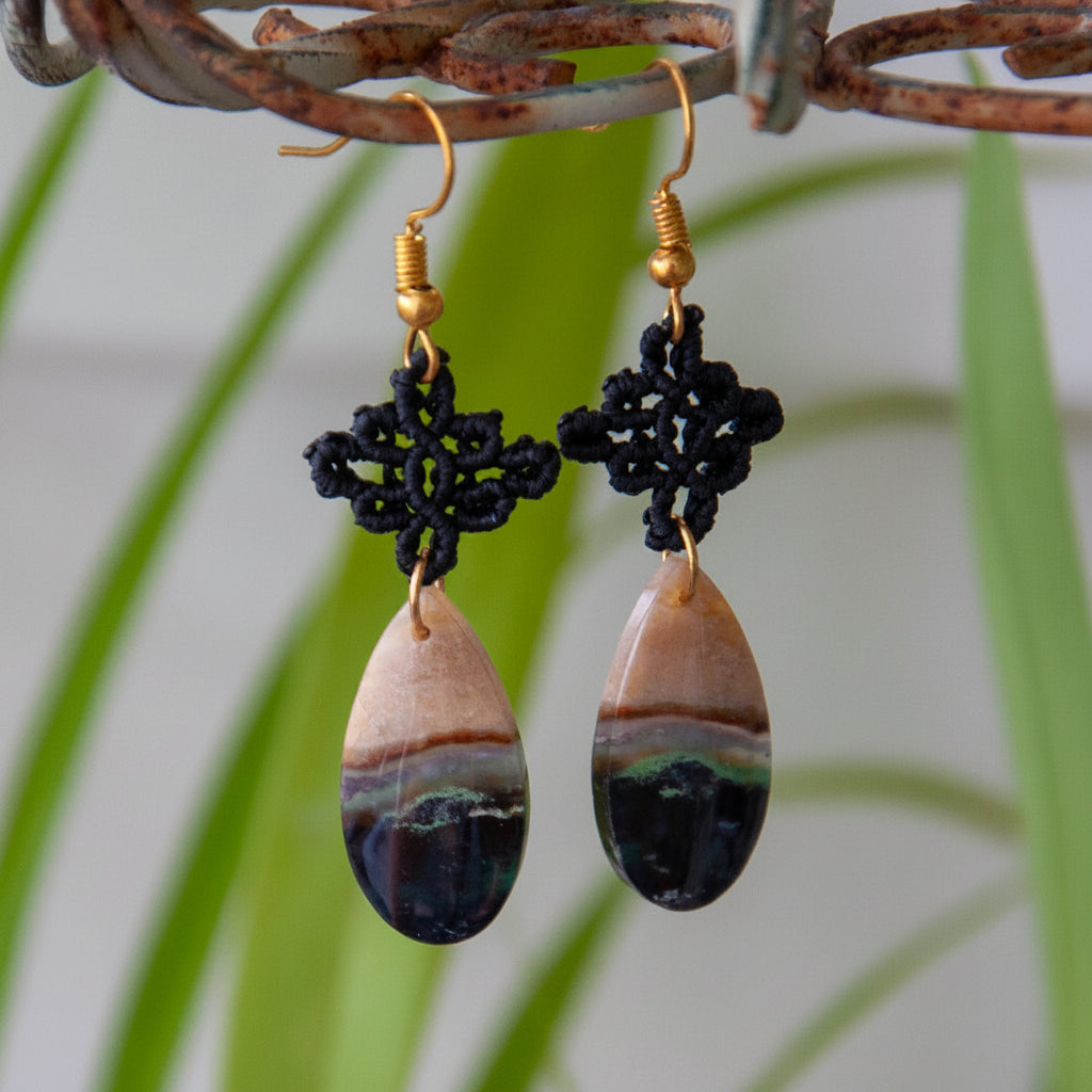 Handmade black macrame drop earrings with diamond-shaped knotwork and green chalcedony teardrop gemstones on gold-tone fish hook wires, hanging from a rustic metal stand with tropical green leaves.