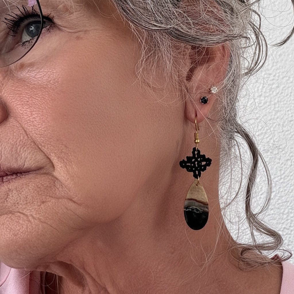 Handmade black macrame drop earring with diamond-shaped knotwork and green chalcedony teardrop gemstone on gold-tone fish hook wire, shown worn on model with silver curly hair and wire-rimmed glasses, pink top.
