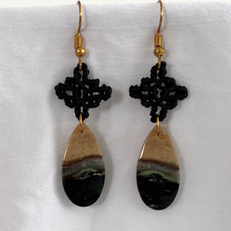Handmade black macrame drop earrings with diamond-shaped knotwork and green chalcedony teardrop gemstones on gold-tone fish hook wires, displayed on draped white fabric.