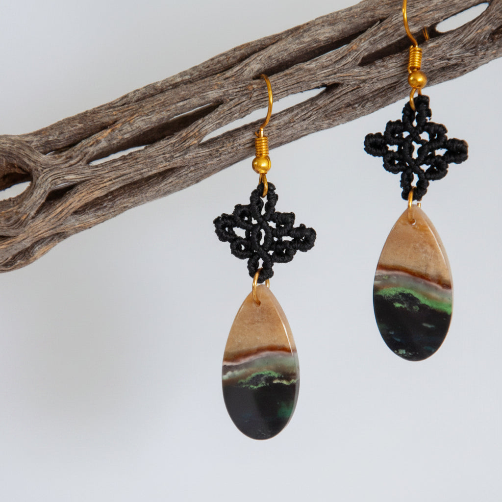 Handmade black macrame drop earrings with diamond-shaped knotwork and green chalcedony teardrop gemstones on gold-tone fish hook wires, hanging from a driftwood branch against a light background.