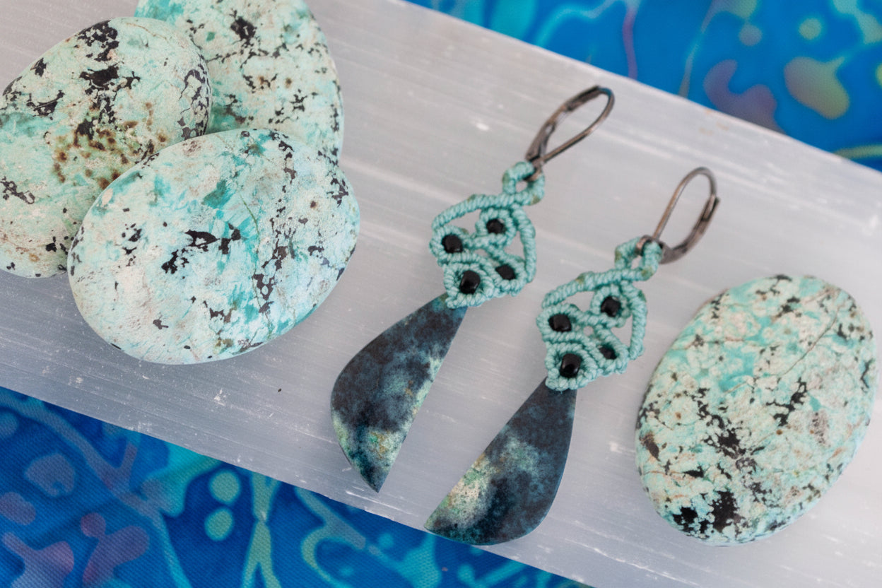 A pair of handcrafted macrame dangle earrings in teal nylon cord with intricate knotwork, black seed bead accents, and hand-selected Bacan stone teardrop pendants in deep teal and charcoal with gunmetal lever back ear wires, displayed on a white selenite slab surrounded by raw Bacan stones on a blue patterned surface.