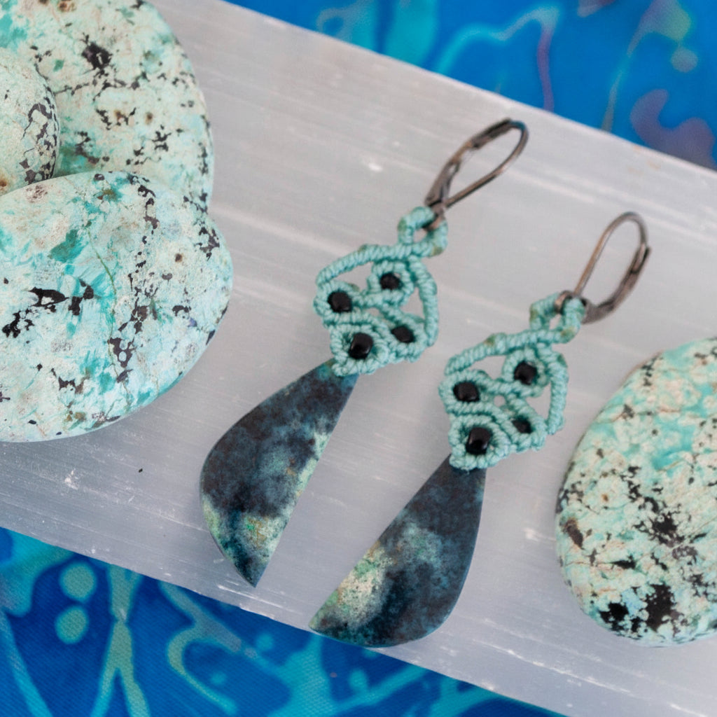 A close-up of a pair of handcrafted macrame dangle earrings in teal nylon cord with intricate knotwork, black seed bead accents, and hand-selected Bacan stone teardrop pendants in deep teal and charcoal with natural mineral patterning, with gunmetal lever back ear wires, displayed on a white selenite slab with raw Bacan stones and a blue patterned background.