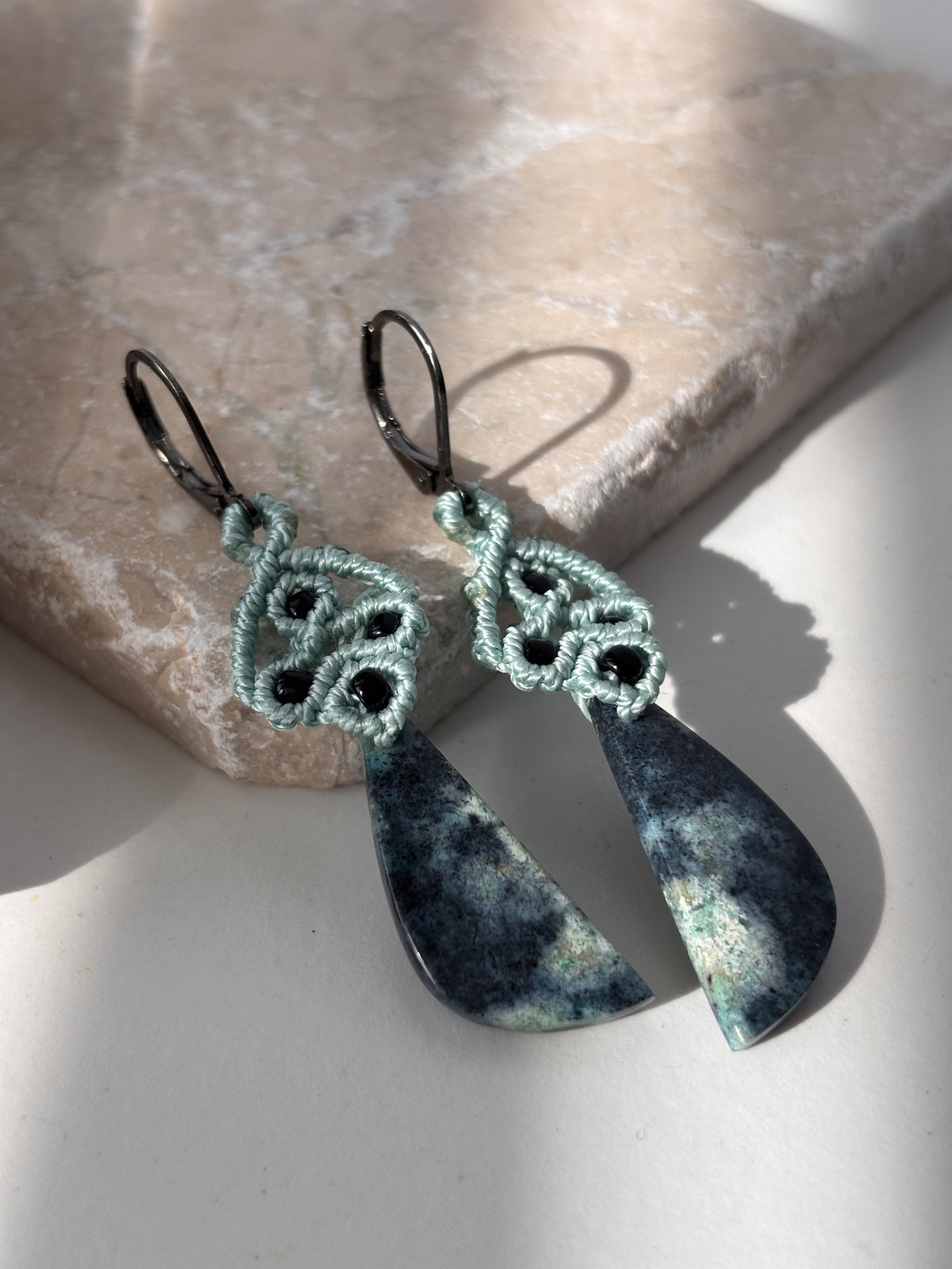 Close-up of teal macrame dangle earrings with black seed bead accents and dark teal and charcoal Bacan stone pendants