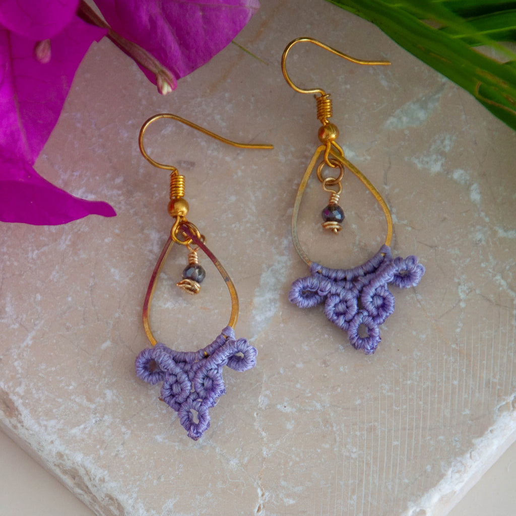 Handmade lavender macrame fan drop earrings with a gold-tone teardrop hoop, brass wire-wrapped faceted purple crystal bead accent, and gold-tone fish hook wires, laid on a travertine tile with magenta bougainvillea and tropical palm leaves.