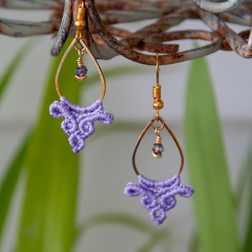 Handmade lavender macrame fan drop earrings with a gold-tone teardrop hoop, brass wire-wrapped faceted purple crystal bead accent, and gold-tone fish hook wires, hanging from a rustic metal stand with tropical green leaves in the background.
