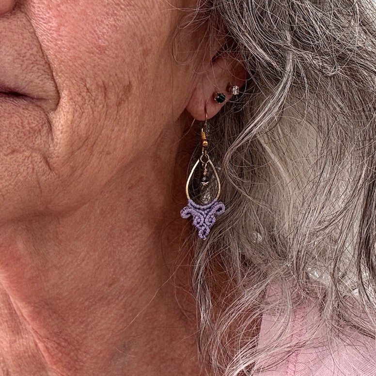 Handmade lavender macrame fan drop earring with gold-tone teardrop hoop and faceted purple crystal bead, shown worn on model with silver hair.