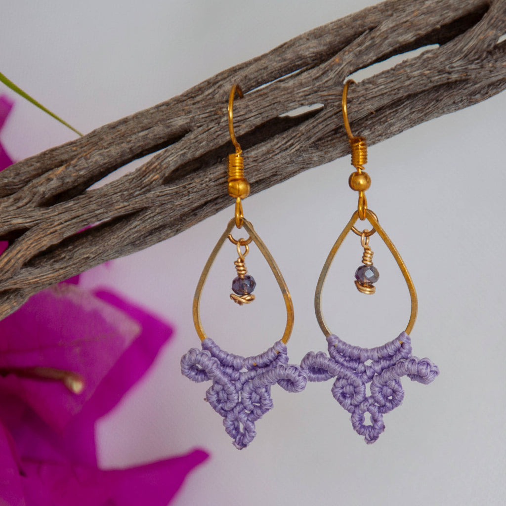 Handmade lavender macrame fan drop earrings with a gold-tone teardrop hoop, brass wire-wrapped faceted purple crystal bead accent, and gold-tone fish hook wires, hanging from a driftwood branch with magenta bougainvillea against a white background.