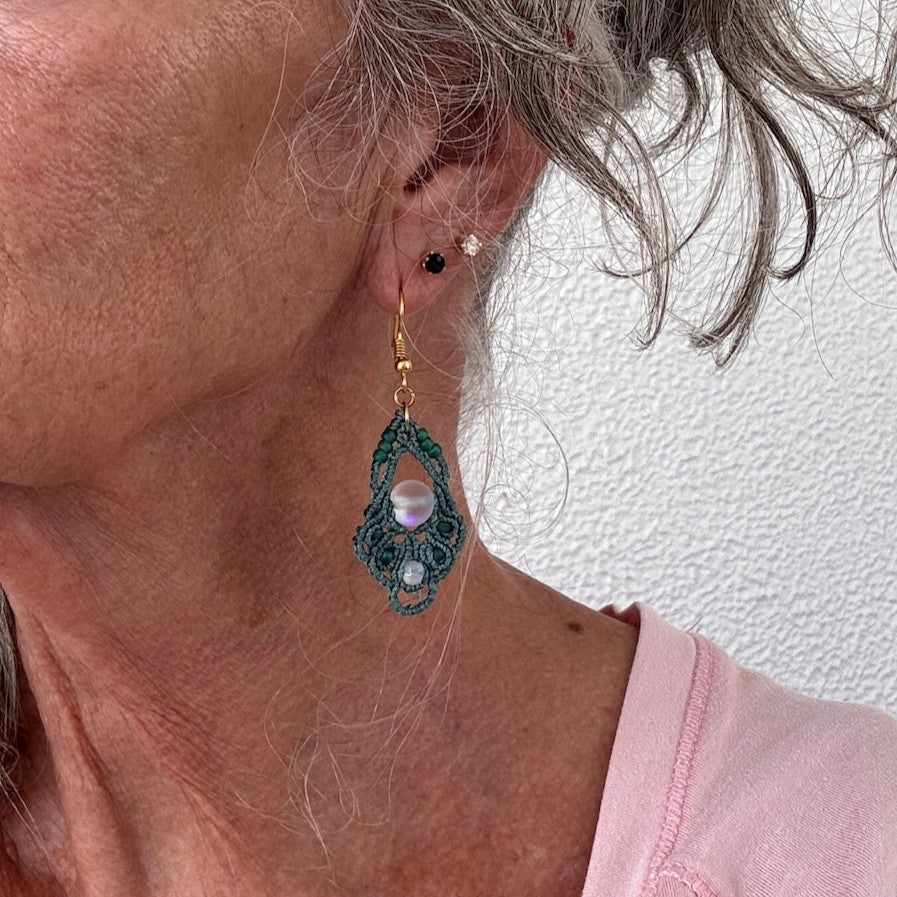 Handmade dark teal macrame diamond drop earring with 8mm iridescent frosted mermaid glass center bead, faceted opalite accent, teal seed beads, and gold-tone fish hook wire shown worn on model with silver hair.