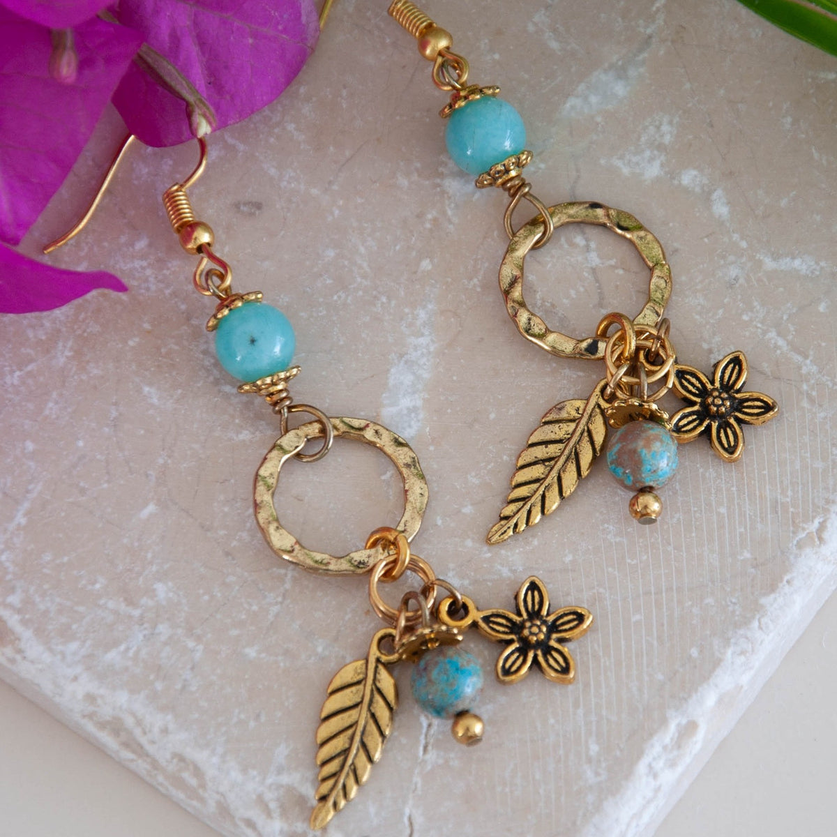 Pair of gold-tone textured ring earrings with amazonite beads, blue calsilica drops, and gold-tone leaf and flower charms, displayed on travertine tile with pink orchid