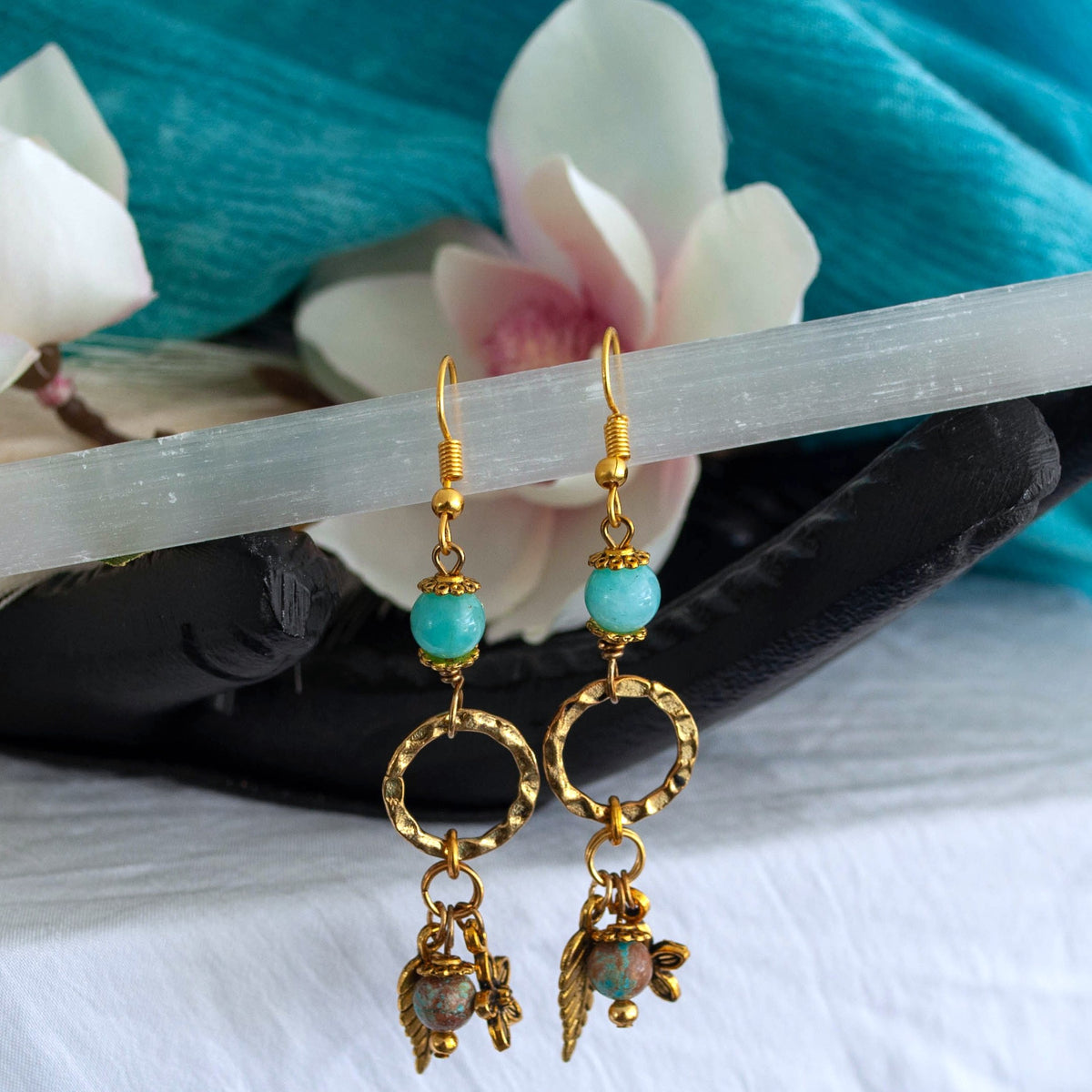 Gold-tone textured ring earrings with amazonite beads and blue calsilica drop charms, displayed on selenite and black stone with magnolia flower and teal linen background