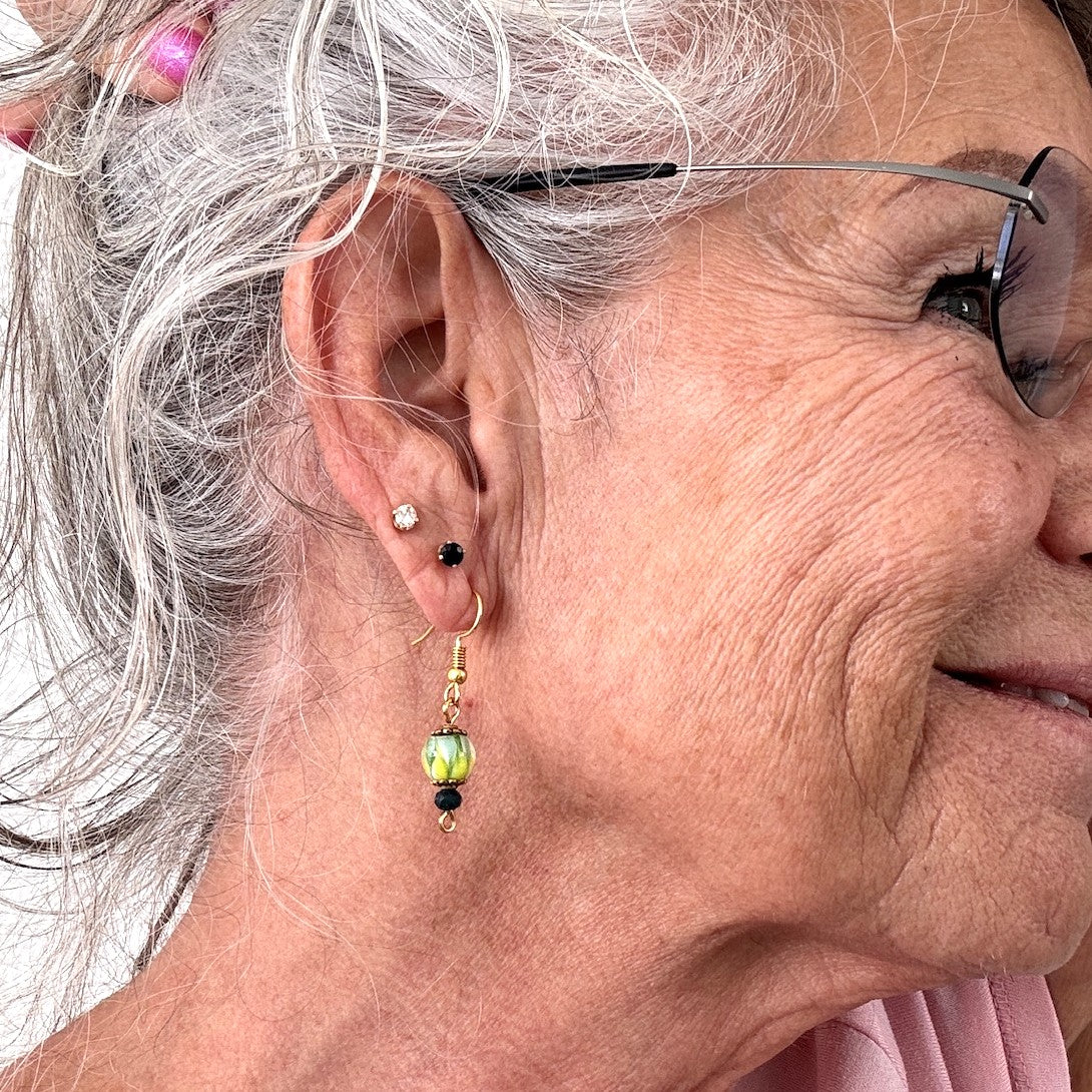 Handmade tropical swirl lampwork glass drop earring with 8mm green, yellow, and white hand-torched lampwork bead, gold-tone daisy spacer beads, 3mm dark green faceted crystal rondelle, and gold-tone fishhook ear wire shown worn on model with silver hair and glasses.