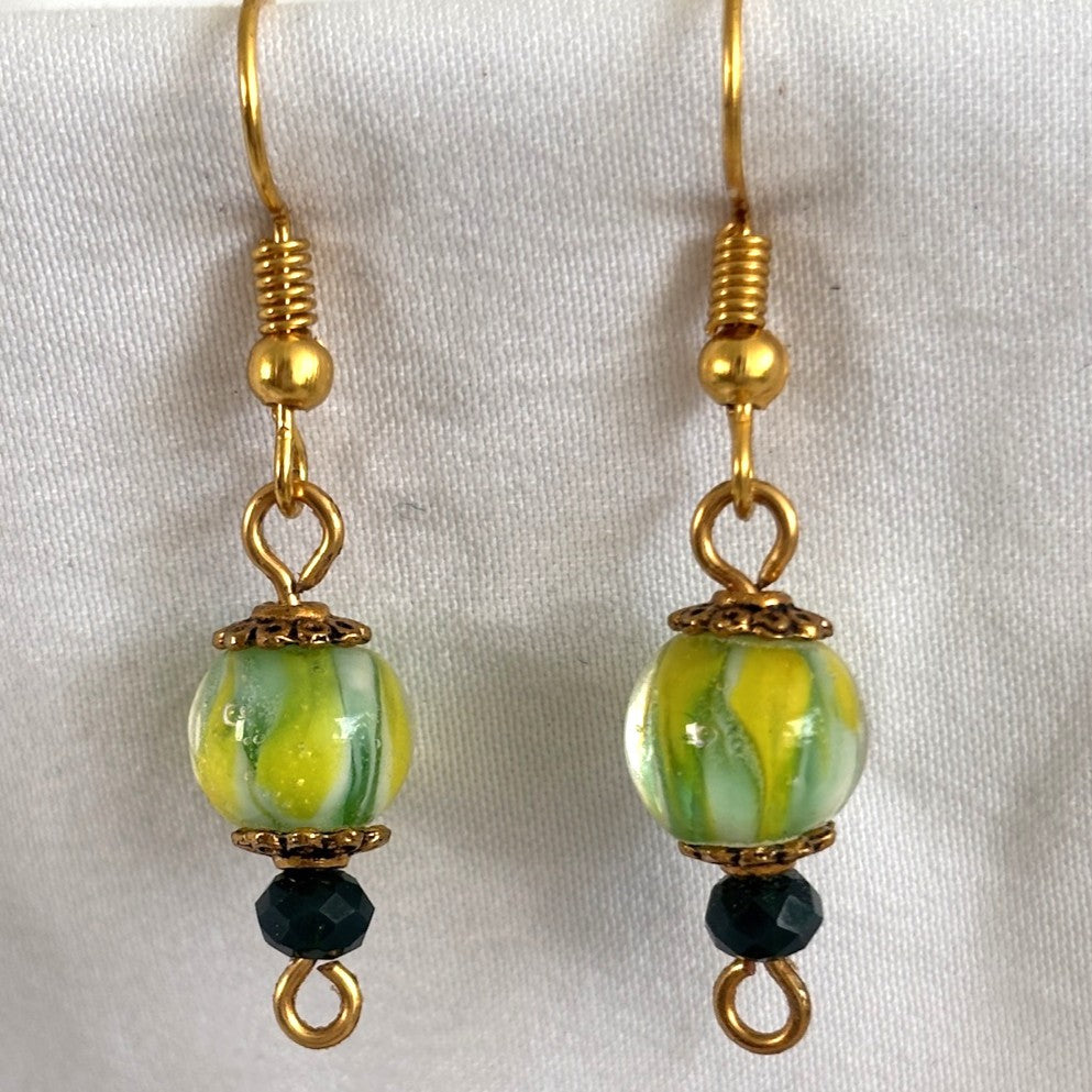 Pair of handmade tropical swirl lampwork glass drop earrings with 8mm green, yellow, and white hand-torched lampwork beads, gold-tone daisy spacer beads, 3mm dark green faceted crystal rondelles, and gold-tone fishhook ear wires, displayed on white fabric background.
