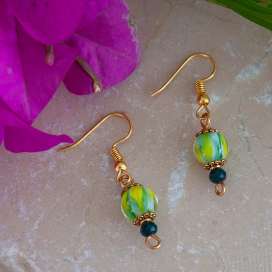 Handmade tropical swirl lampwork glass drop earrings with 8mm green, yellow, and white lampwork bead, gold-tone daisy spacer beads, 3mm dark green faceted crystal rondelle, and gold-tone fishhook ear wires, laid on travertine tile with magenta orchid and tropical palm leaves.