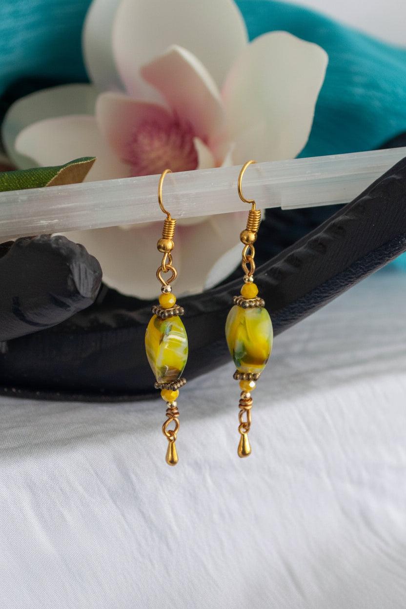 Handmade chartreuse twisted rectangle lampwork glass drop earrings with square gold-tone spacer beads, 3mm opaque yellow crystal, gold-tone teardrop dangle, and fishhook ear wires, hanging from black driftwood with a white magnolia flower and teal background.