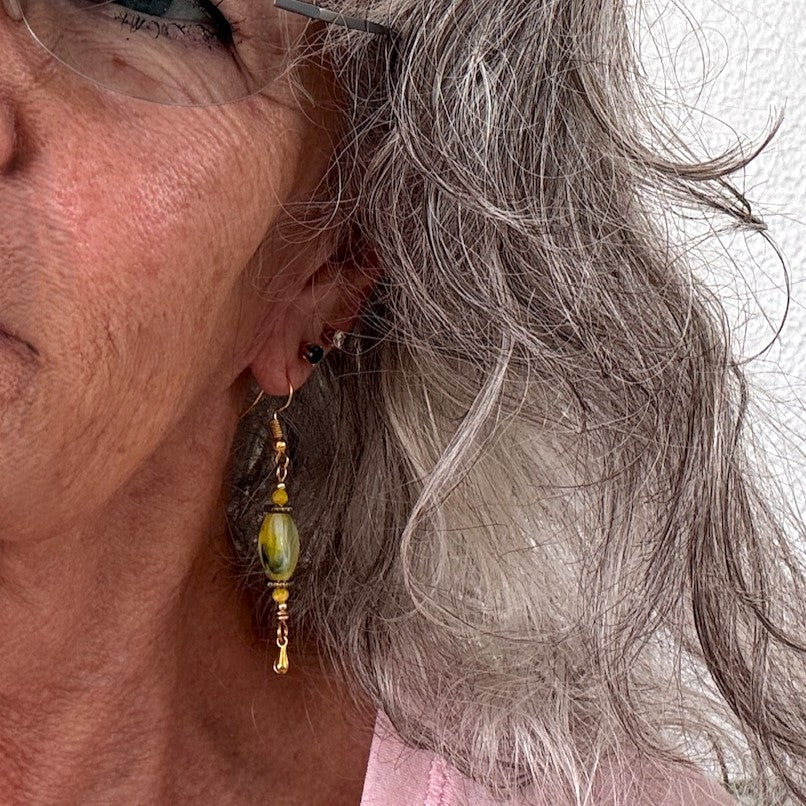Handmade chartreuse twisted rectangle lampwork glass drop earring with square gold-tone spacer beads, 3mm opaque yellow crystal, gold-tone teardrop dangle, and gold-tone fishhook ear wire shown worn on model with silver hair against white background.
