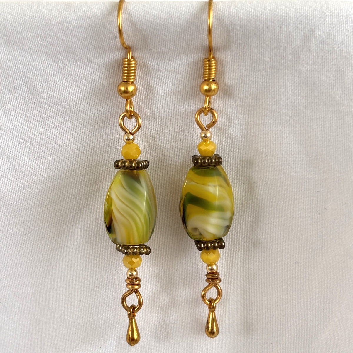 Pair of handmade chartreuse twisted rectangle lampwork glass drop earrings with square gold-tone spacer beads, 3mm opaque faceted yellow crystal beads, 3mm gold-tone teardrop dangles, and gold-tone fishhook ear wires, displayed on white fabric background.